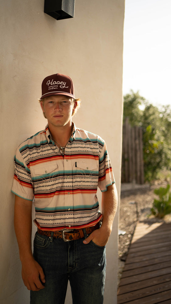 male model posing against a wall wearing medium wash jeans, red/blue/white striped sol shirt, and maroon Hooey hat