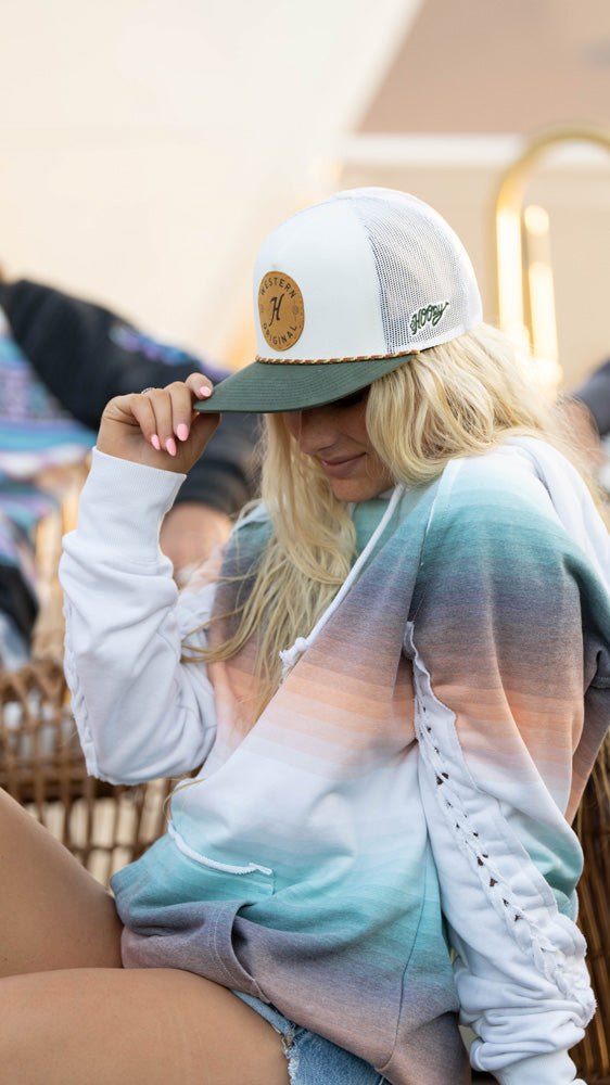close up of female model wearing a white and grey hooey hat with pink/blue/purple/white hooey hoody posing with a hand on her hat  while seated in an outdoor setting
