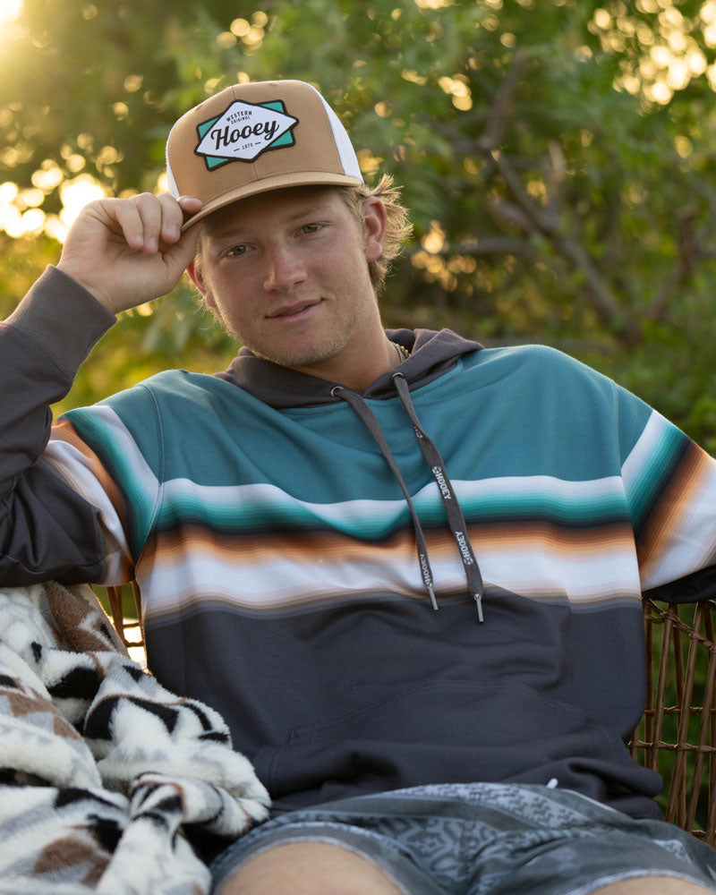 young male model wearing grey and white board shorts, grey/turquoise/orange striped hoody with tan and white Hooey hat posing in outdoor setting with hand on hat