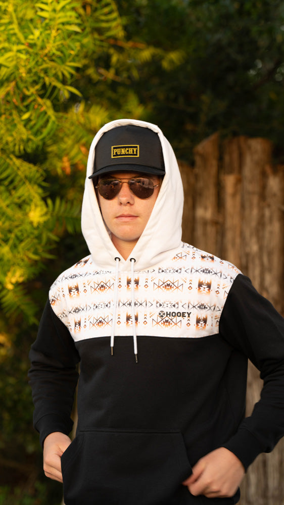 male model wearing black and white Aztec hooey hoodie with black and gold hat and sun glasses posing in outdoor setting with hands on hem of hoody