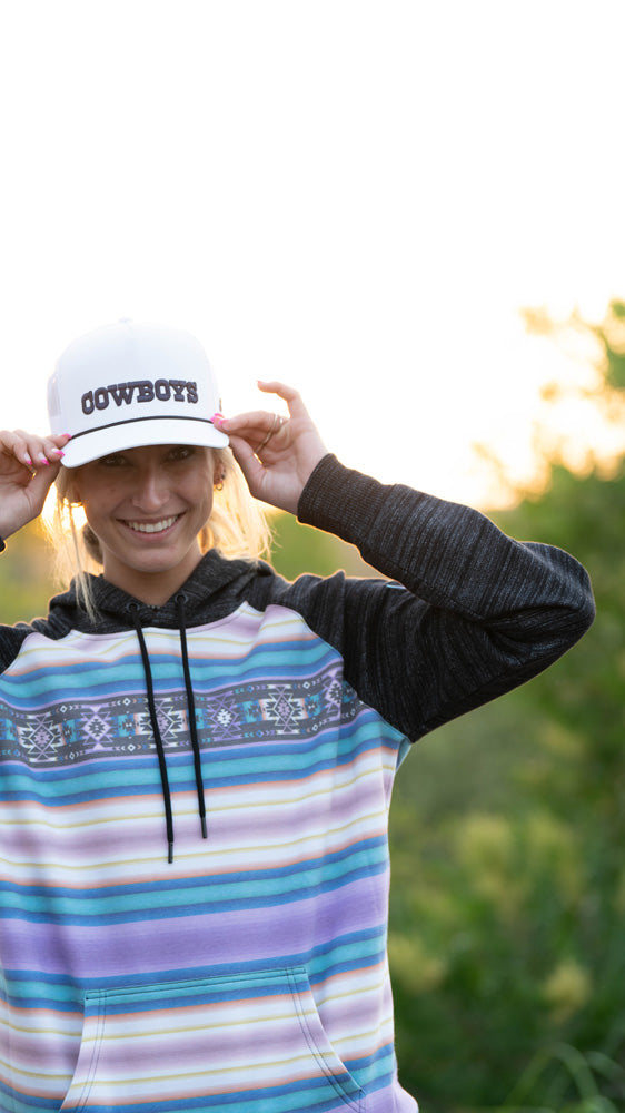 female model wearing white and navy Dallas Cowboys x Hooey hat with purple/blue/white serape hoody posing with both hands on hat in outdoor setting