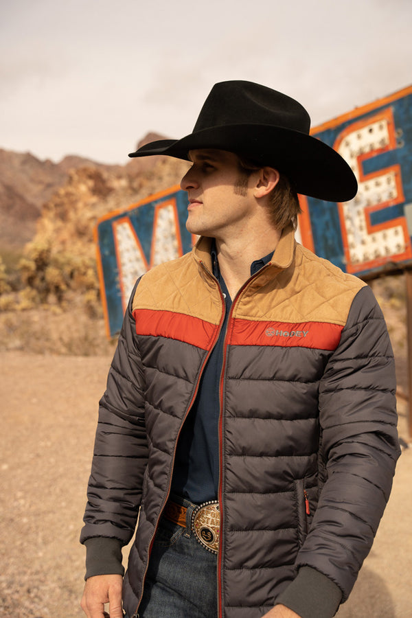 male model wearing grey, tan, and red puffer jacket, blue jeans, and black felt cowboy hat posed in desert junk yard