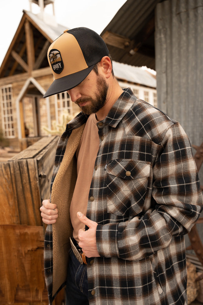 Profile photo of male model wearing dark washed blue jeans and belt buckle gray and brown plaid Shacket brown T-shirt tan and black Cheyenne hooey hat