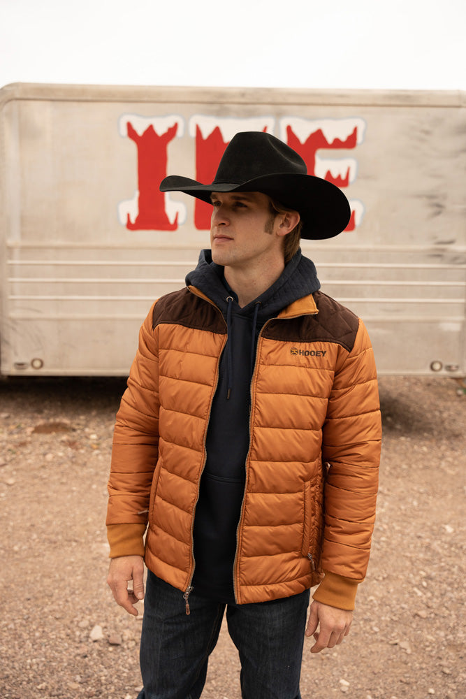 Male model rain dark washed blue jeans brown and tan puffer jacket black hoodie black felt cowboy hat posing in front of old ice trailer