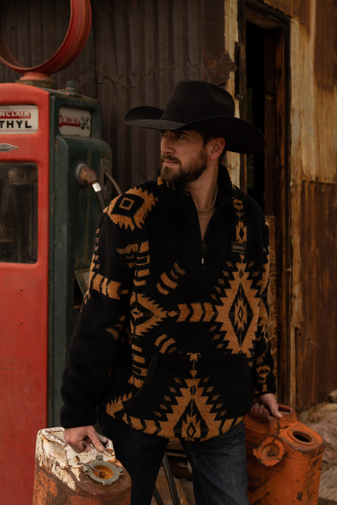 male model wearing the Hooey fleece pullover in black with Aztec pattern in tan, felt cowboy hat, carrying rusted gas cans