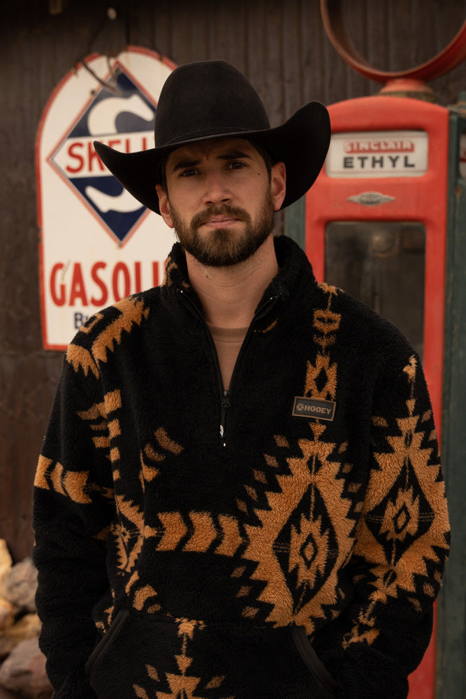 male model wearing the Hooey fleece pullover in black with Aztec pattern in tan, black felt cowboy hat in front of a rustic gas pump