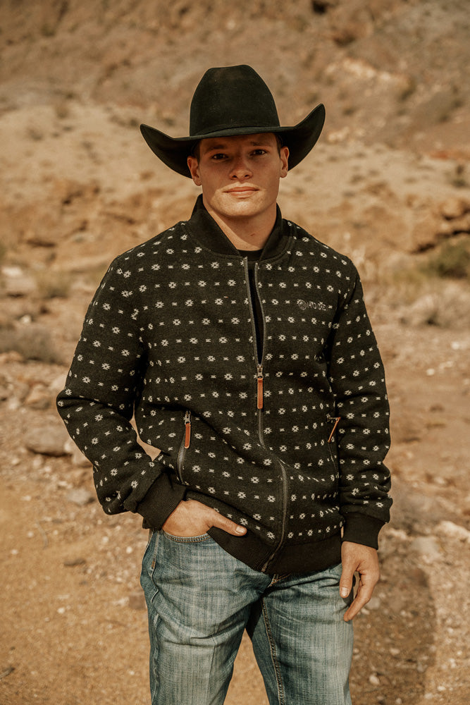 Male model wearing medium wash blue jeans black jacket with micro pattern black felt cowboy hat posing in desert