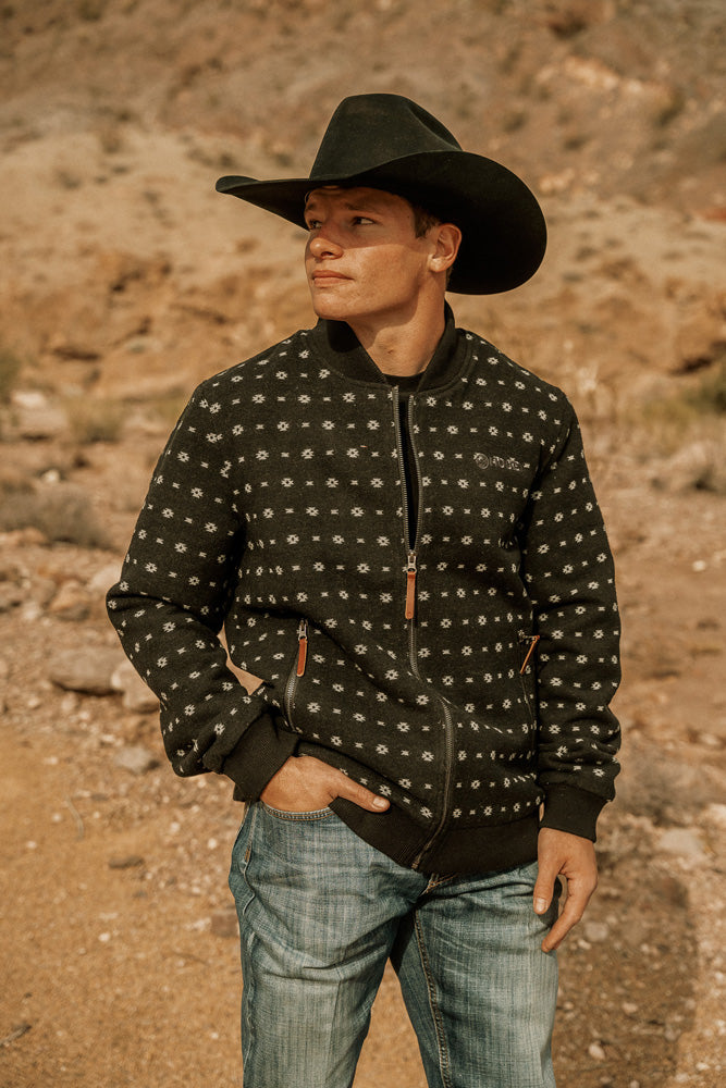 Male model posing in desert setting where in light wash jeans black zipper jacket with micro pattern and black felt cowboy hat