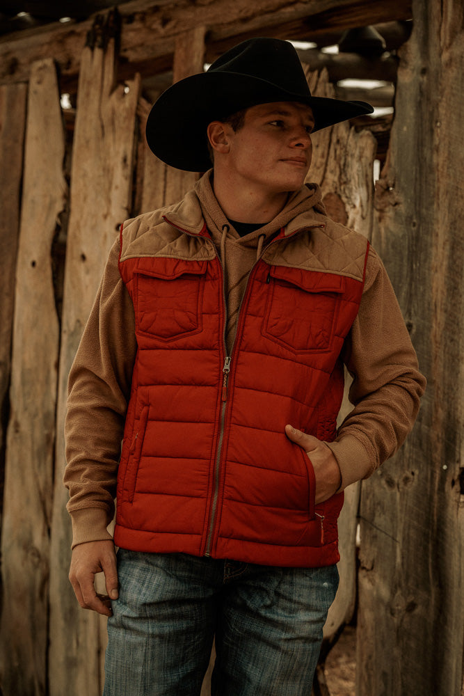 Male model wearing light wash blue jeans red and brown puffer jacket and black cowboy hat posing in front of wooden structure