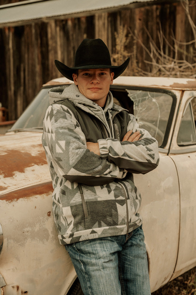 Male model wearing light washed blue jeans gray and white Aztec print jacket and black felt cowboy hat leaning against rusty car