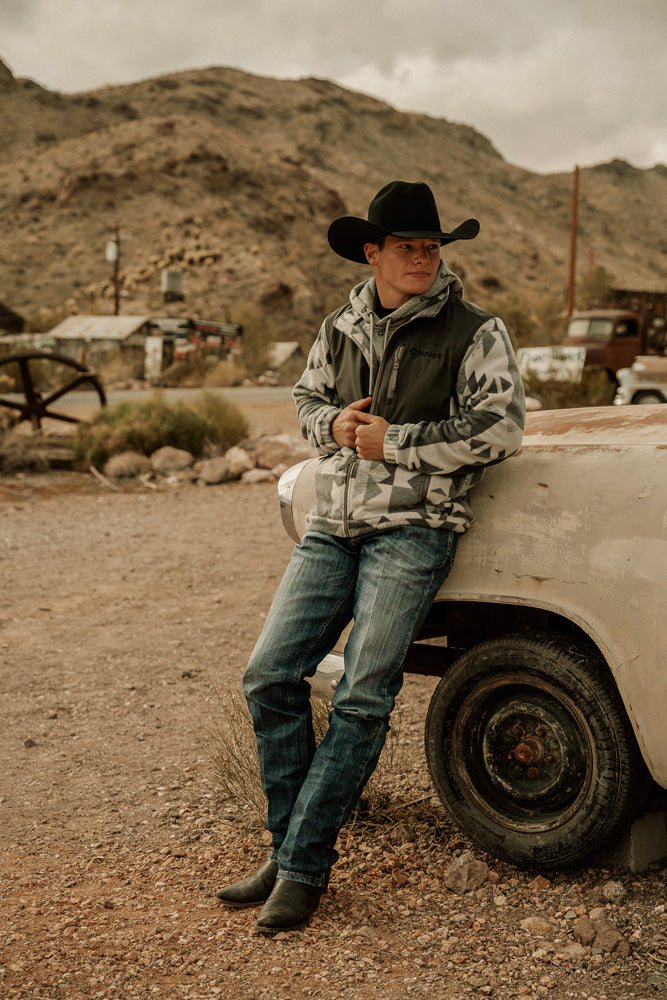 Male model wearing black cowboy boots medium wash blue jeans gray and white hooey Jacket Black Felt Cowboy Hat Propped Against Rusted Old Car