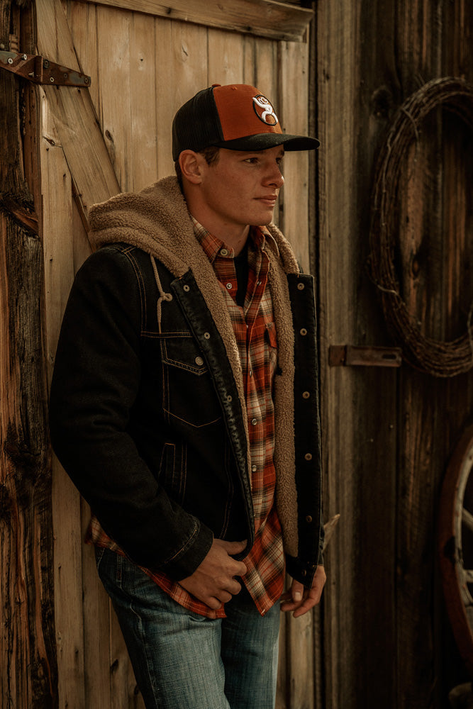 Male model wearing white wash blue jeans orange and brown plaid button down Shacket with dark wash denim jacket featuring brown fleece lining and an orange and black roughy hat