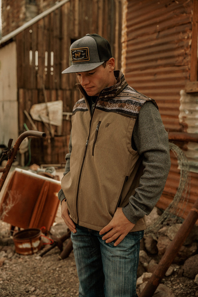 Male model wearing medium wash blue jeans gray long sleeve shirt tan Hooey vest with brown and gray Aztec print on shoulders and collar and a gray and black rank stock rodeo company hat