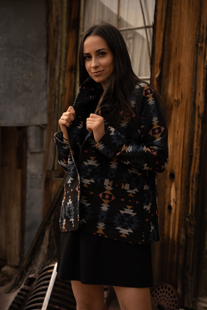 female model wearing black bamboo dress with black jacket featuring orange, blue, and white Aztec pattern posed in front of wooden building