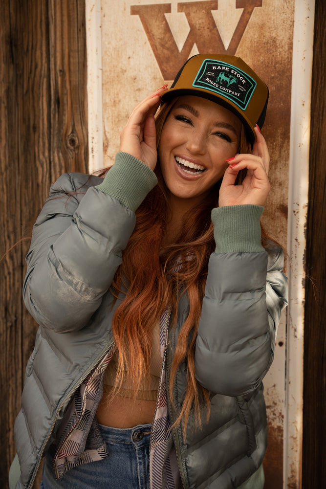female model wearing medium wash jeans, olive puffer jacket, and brown rank stock rodeo company hat posing against a rustic metal sign