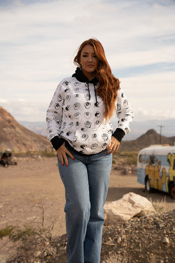 red haired, female model wearing the vaquera white hoody with black cowboy hat pattern, black hod, and cuffs with jeans in desert setting