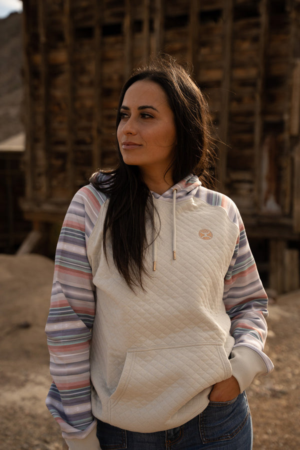 female model wearing the summit cream hoody with pink, grey, orange aztec pattern on pocket, sleeves, and hood with jeans in desert setting