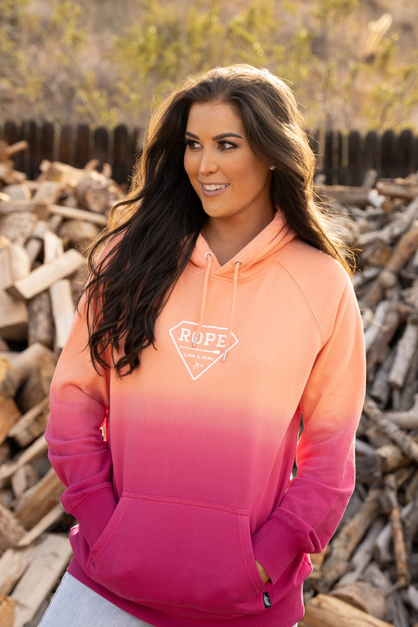 lifestyle image of a brunet female model wearing the ROPE LIKE A GIRL pink and orange ombre hoody standing infront of a pile of fire wood in outdoor setting
