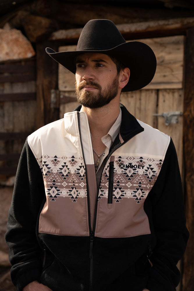 Male model with facial hair wearing black felt cowboy hat black pull over with brown and white Aztec print on the upper half of the torso