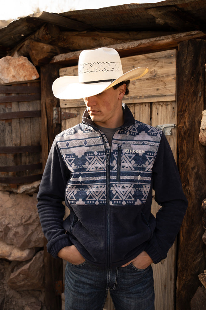 Male model wearing dark wash bluejeans navy fleece pull over with Aztec pattern and straw cowboy hat posing in front of rustic wooden structure