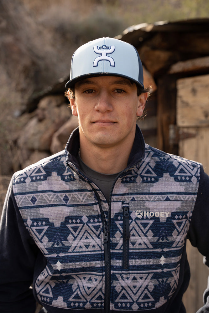 Close-up photo of male model wearing blue hooey hat with white logo and navy fleece an Aztec pull over
