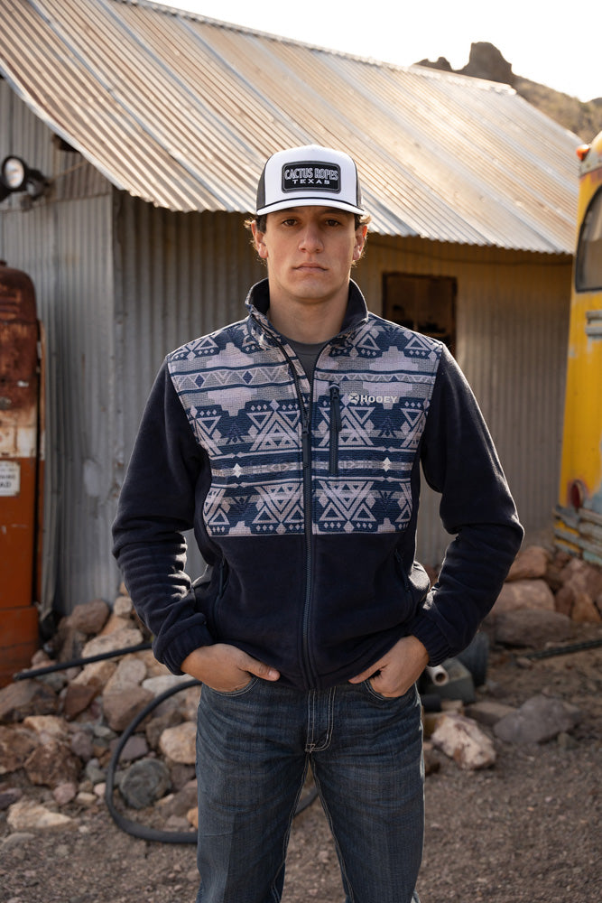Male model wearing dark wash blue jeans navy fleece pull over with navy and light gray Aztec print across the collar and chest black and white cactus ropes Texas cap posing in rustic desert setting