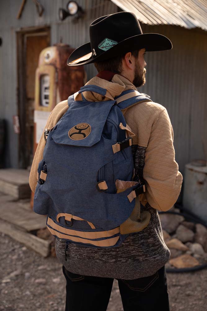 Male model sporting the Mule denim backpack with brown strap features posing infront of a rusty metal building