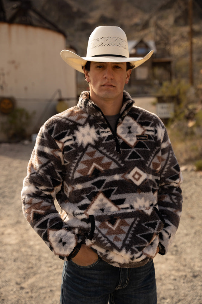 male model posing in dark wash jeans, grey and brown aztec pullover and straw cowboy hat in desert setting