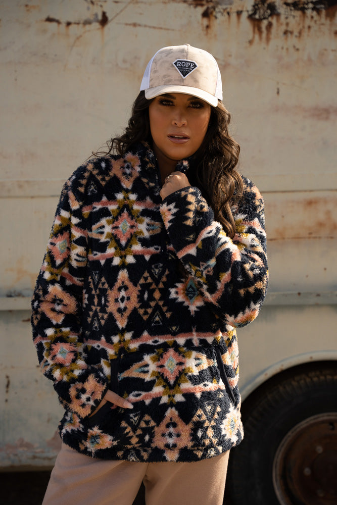 female model wearing tan pants, black and multi colored Aztec pattern pullover with tan and white RLAG cap