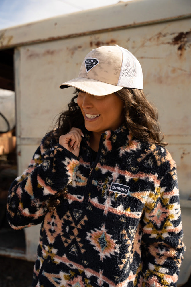 female model wearing black/pink/gold/tuquoise Aztec print pullover with tan and white RLAG cap posed in front of rusted out box truck