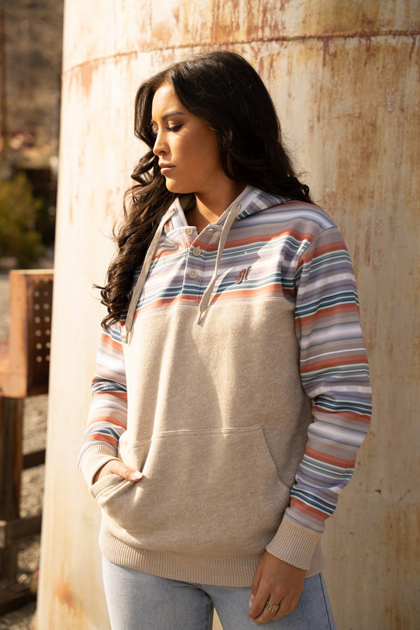 female model sporting the Jimmy heathered tan hoody with grey, green, and tan stripe pattern on sleeves, collar, and hood with jeans in an outdoor setting