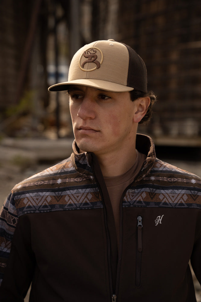 male model wearing tan and brown hooey roughy cap, with dark brown and multi pattern zipper jacket