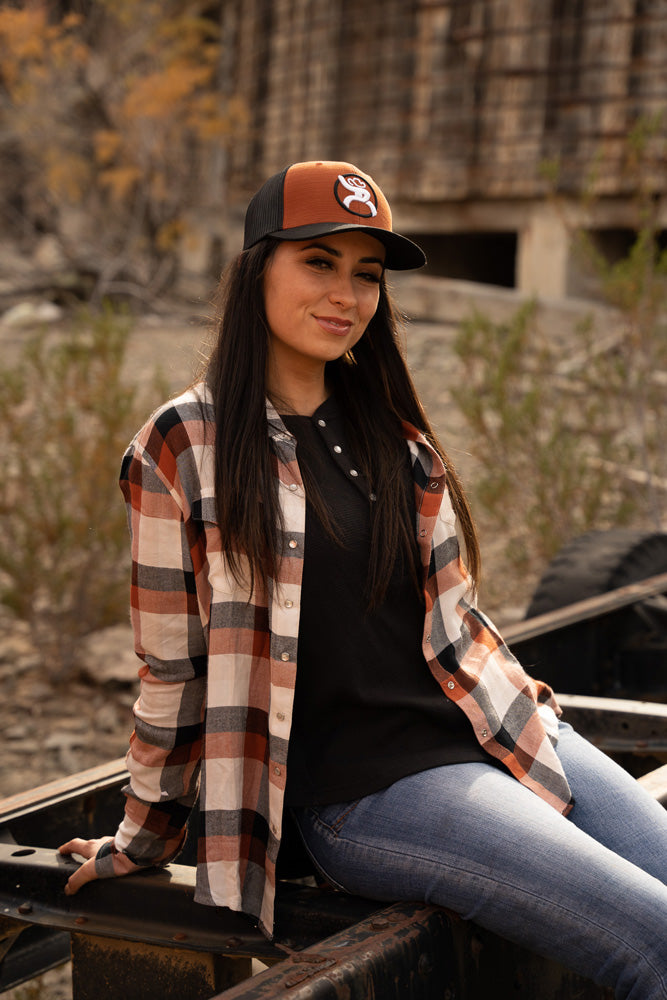 female model wearing orange and black hat with black and white hooey logo, orange, cream, and grey plaid shacket, black henley and blue jeans seated on a trailer chasey 