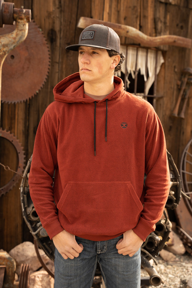 male model wearing a grey hat, jeans, and the shadow red hoody with Hooey logo on back infront of a rustic garage 