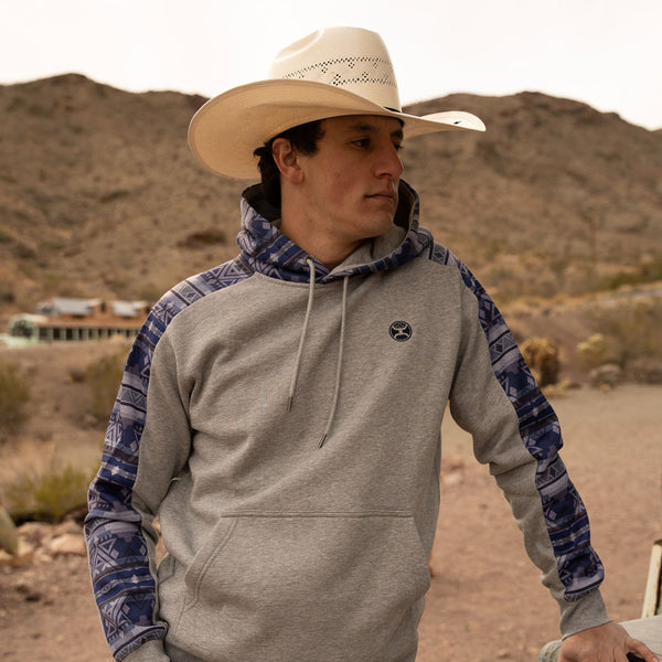 male model sporting the Canyon grey hoody with navy blue aztec pattern on sleeves and hood, wearing a straw cowboy hat in a mountainous setting. 