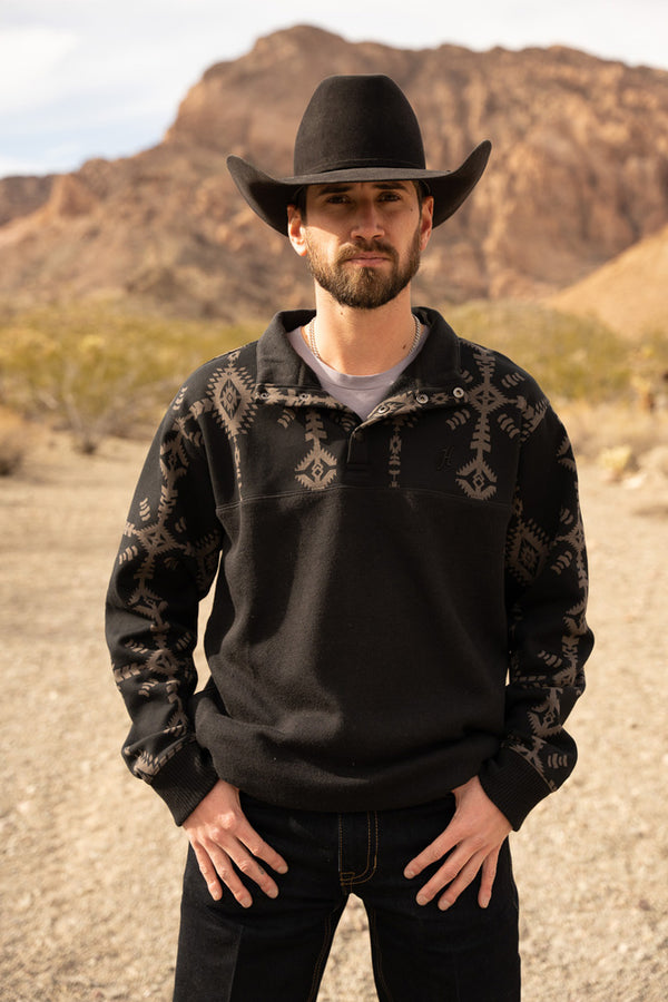 male model wearing the Stevie black pullover with black and grey Aztec pattern on collar and sleeves, black jeans, and felt cowboy hat in mountainous setting