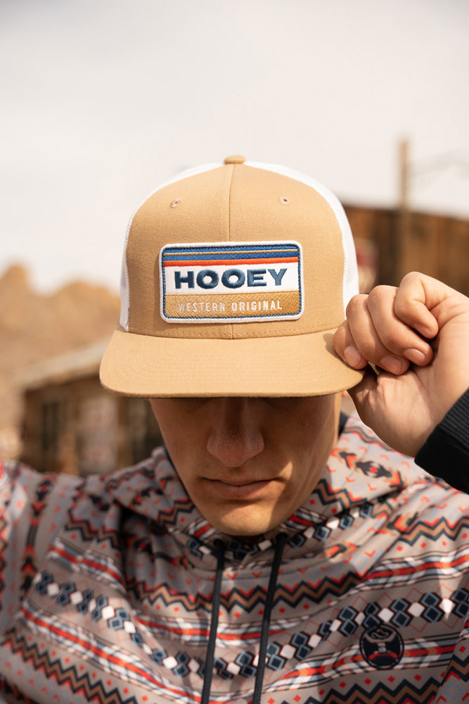 male model wearing multi colored and pattern hoody with tan and white hooey hat pulled over eyes with left hand