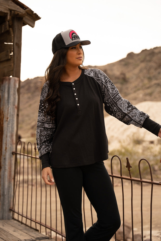 female model posing on the balcony wearing black pants, black henley with black and white bandana pattern on sleeves, black and white hat with red patch