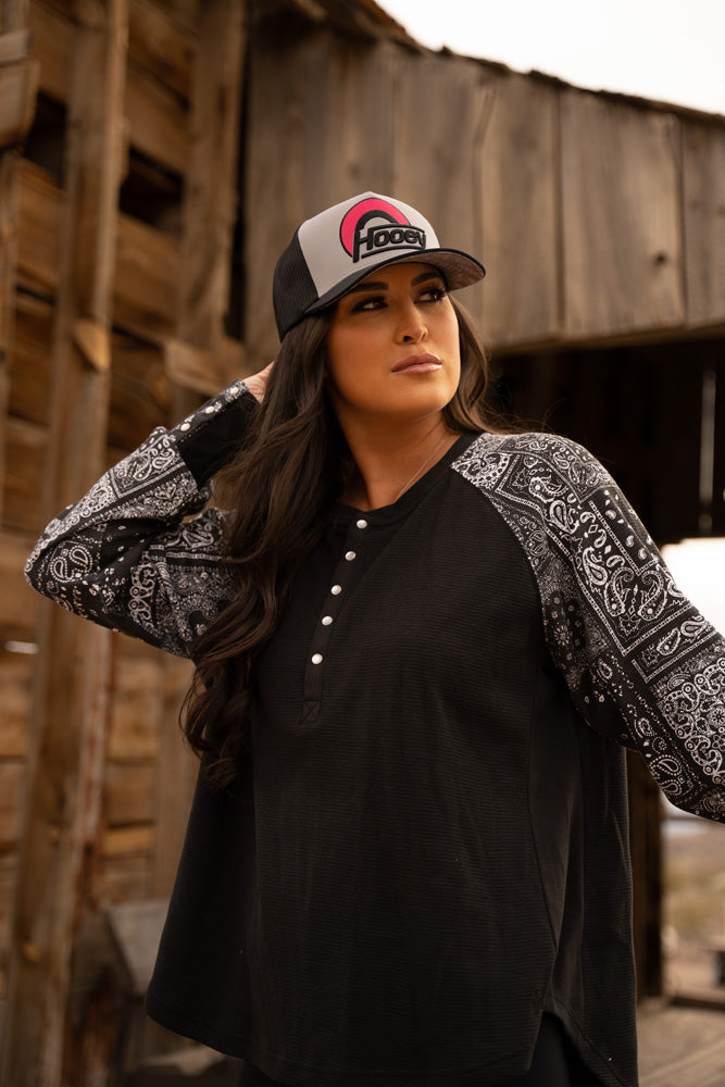 close up of female model wearing black and white hat with red and black Hooey patch, black henley with black and white bandana pattern sleeves posing in front of wooden building