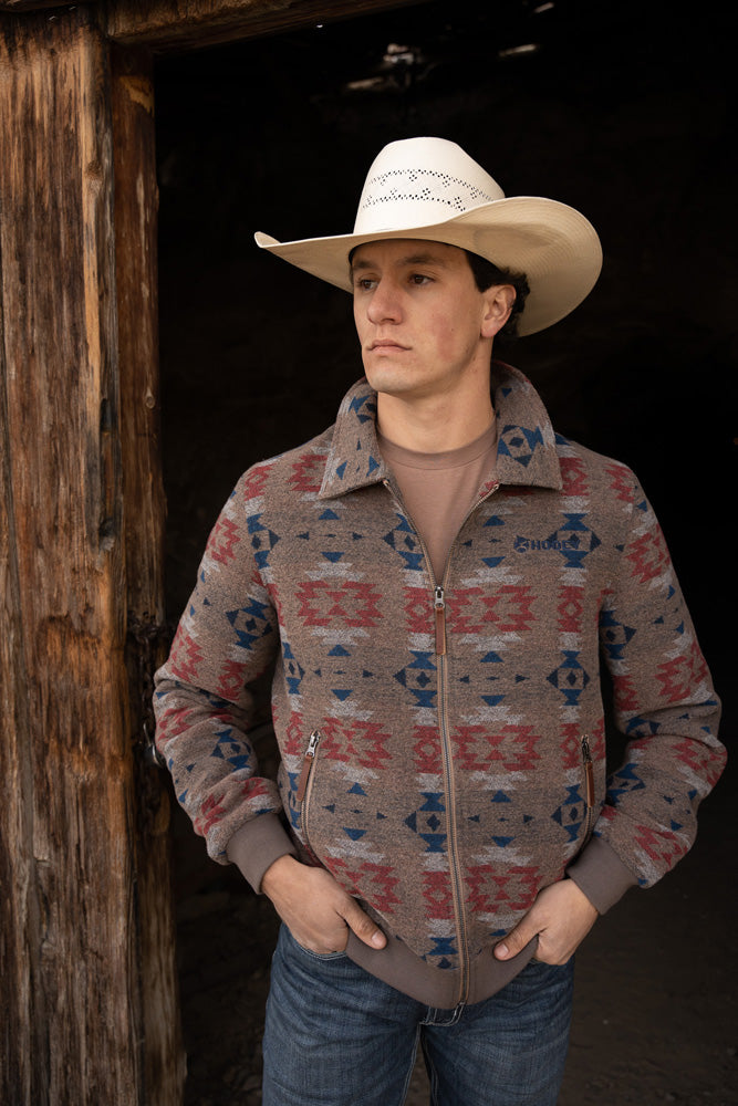 male model wearing blue jeans, brown zipper jacket with red and blue Aztec patter and straw cowboy hat posing in doorway