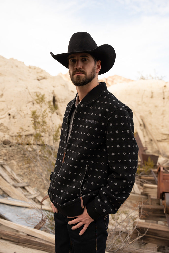 male model wearing black pants, black jacket with micro pattern, black felt cowboy hat posed in junk yard