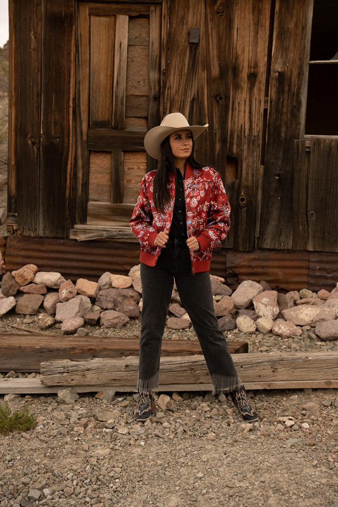 dark haired, female model posing in red, floral bomber jacket, dark pants, and western boots in rusting outdoor setting