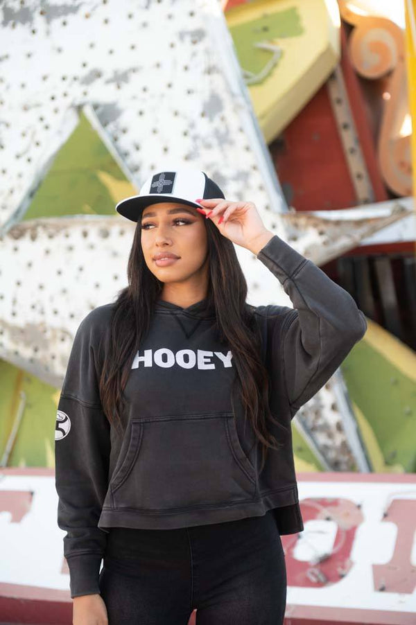 female model wearing the Roomy black hoody with white logo, black and white hat, and black pants posed  infront of signs