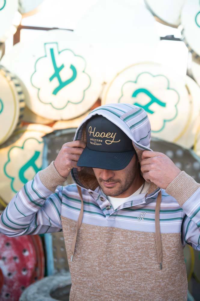 male model sporting the Jimmy heathered tan hoody with grey, green, and tan stripe pattern on sleeves, collar, and hood with hood up, black and gold Hooey hat infront of a pile of old signs