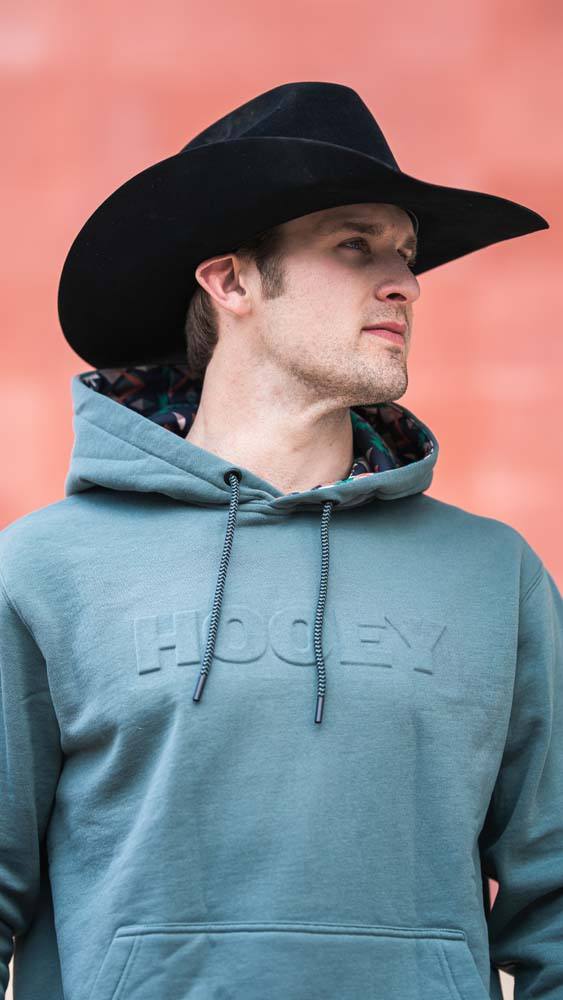 male model wearing a felt cowboy hat and green hoody with multi colored aztec pattern in the hood lining