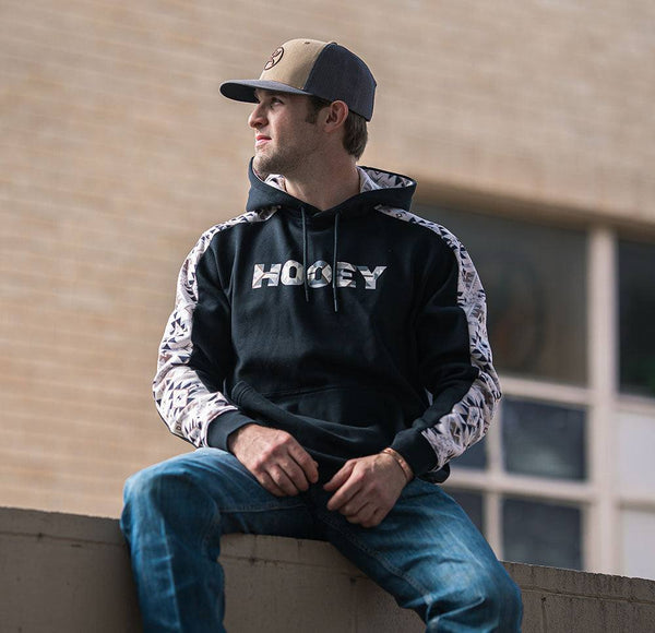 model wearing the Canyon black hoody with white and tan aztec pattern on sleeve and lining sitting on a wall