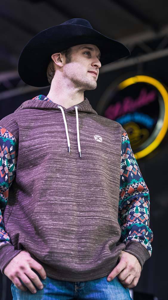 male model in felt cowboy hat and jeans modeling the summit heather brown hoodie with multi collored aztec patten on sleeve, posing infront of a neon sign