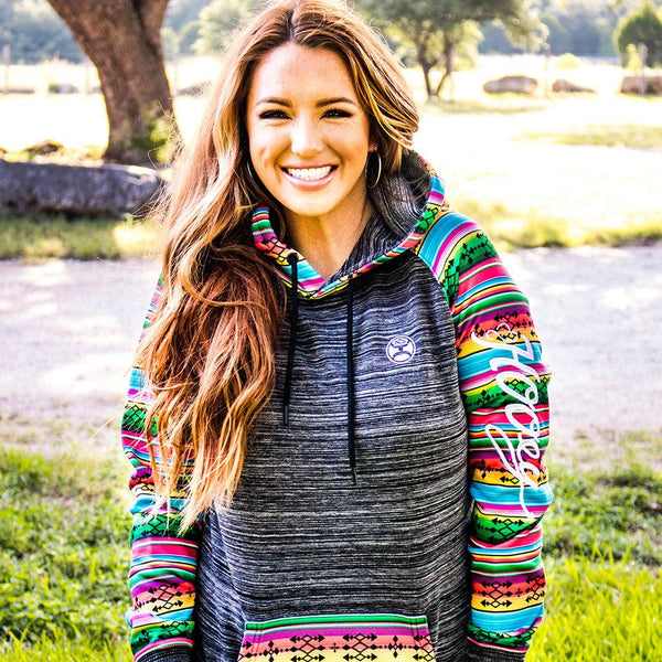 close up of red headed female model wearing the saldo black space dye hody with green blue orange, purple, white stripe pattern on sleeves, pocket, and hood in outdoor setting