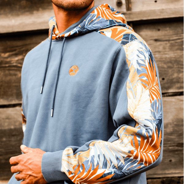 close up of male model wearing the Oasis blue hoody with gold, white, blue, orange leaf pattern on sleeves and hood infront of rustic wood barn