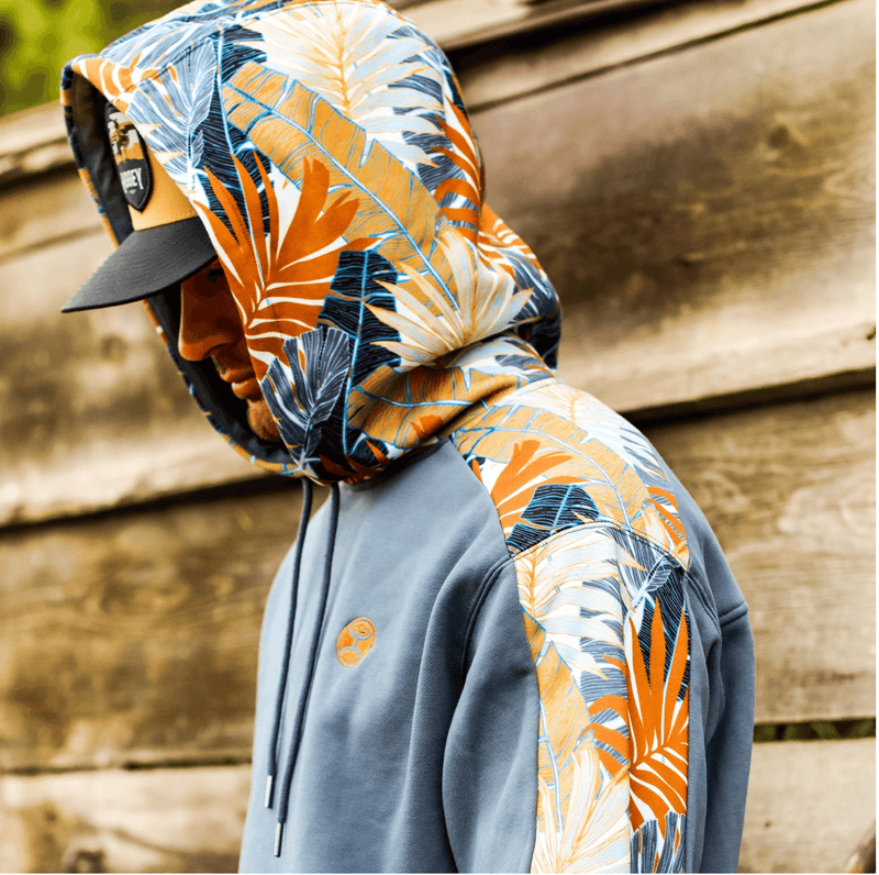 close up of male model wearing the Oasis blue hoody with gold, white, blue, orange leaf pattern on sleeves and hood with hood up, tan and black cheyenne hat infront of rustic wood barn